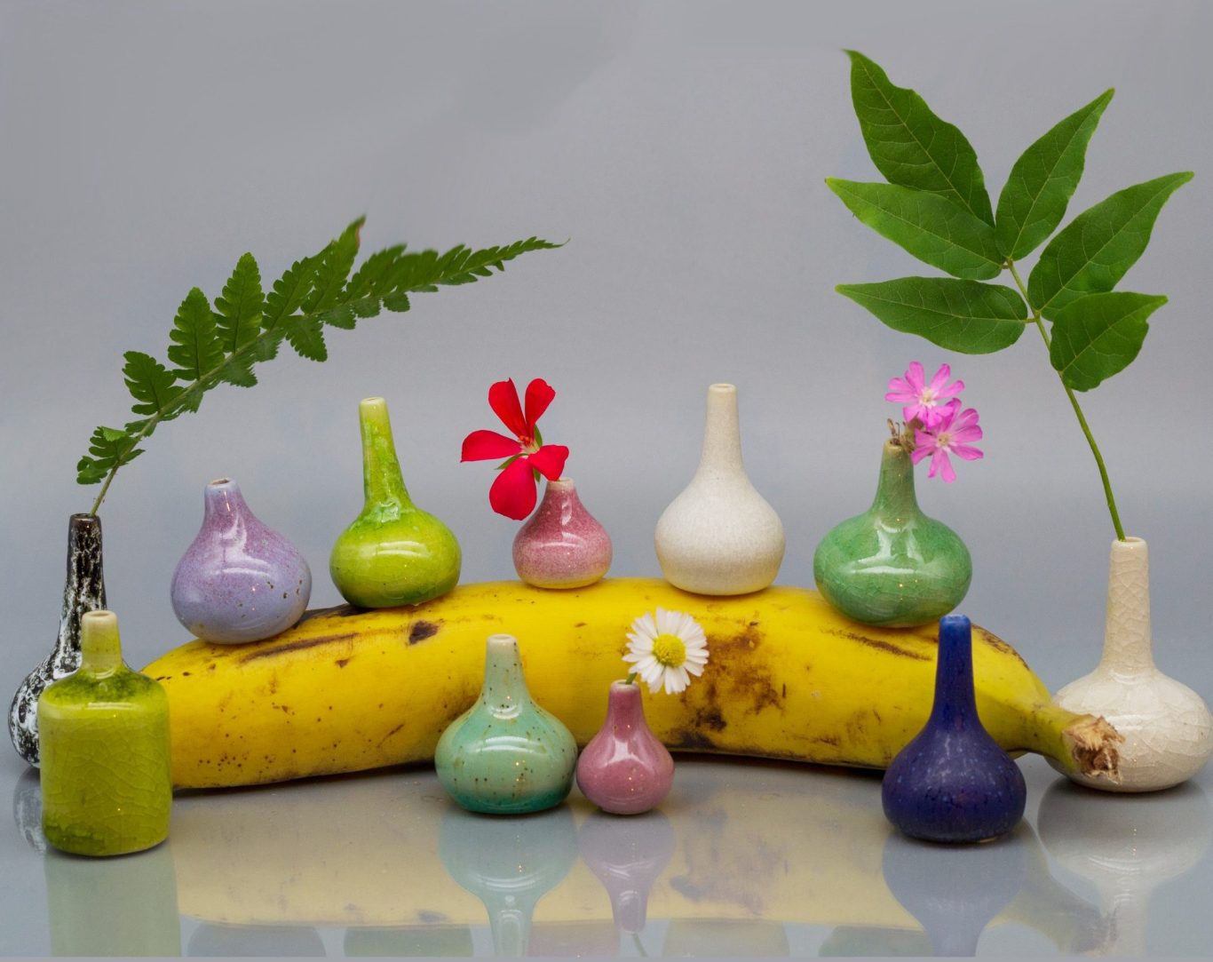 Banana for scale Små färgglada vaser bland en banan och blommor.